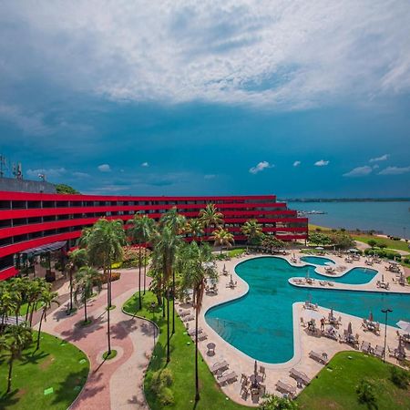 Royal Tulip Brasilia Alvorada Hotel Exterior foto The photo shows a modern hotel building with a striking red exterior, surrounded by lush greenery. In the foreground, there is a large, creatively shaped swimming pool, with lounge chairs arranged around it. The backdrop features a body of water, pos