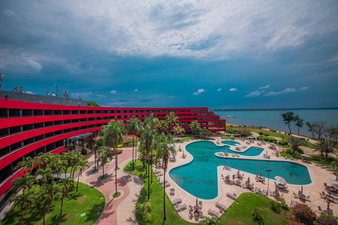 Royal Tulip Brasilia Alvorada Hotel Exterior foto The photo shows a modern hotel building with a striking red exterior, surrounded by lush greenery. In the foreground, there is a large, creatively shaped swimming pool, with lounge chairs arranged around it. The backdrop features a body of water, pos