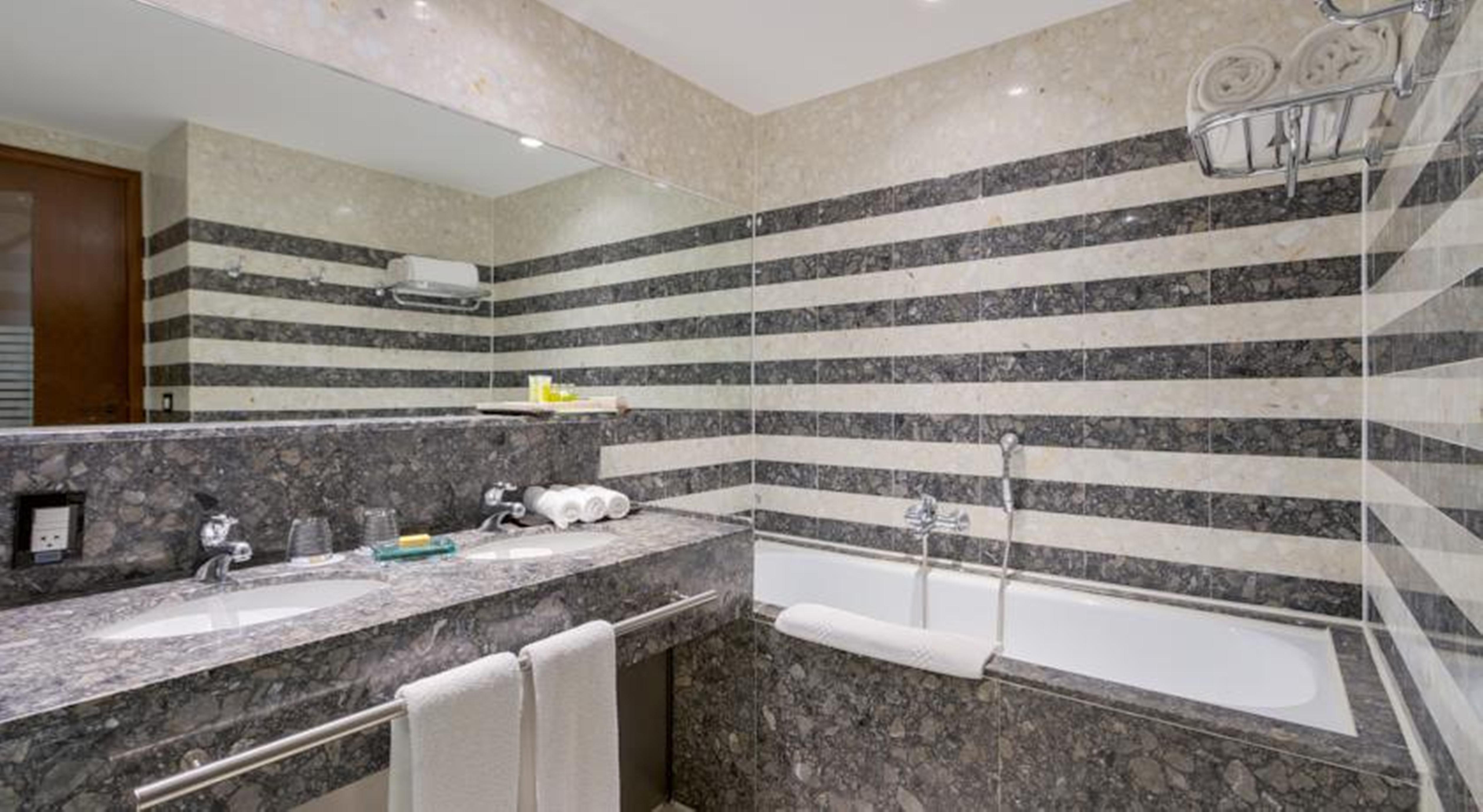 Royal Tulip Brasilia Alvorada Hotel Exterior foto The photo shows a modern bathroom featuring a combination of marble and stone finishes. The walls are decorated with horizontal stripes in various shades of gray and cream. There is a bathtub with a white exterior, and a showerhead is mounted on the 