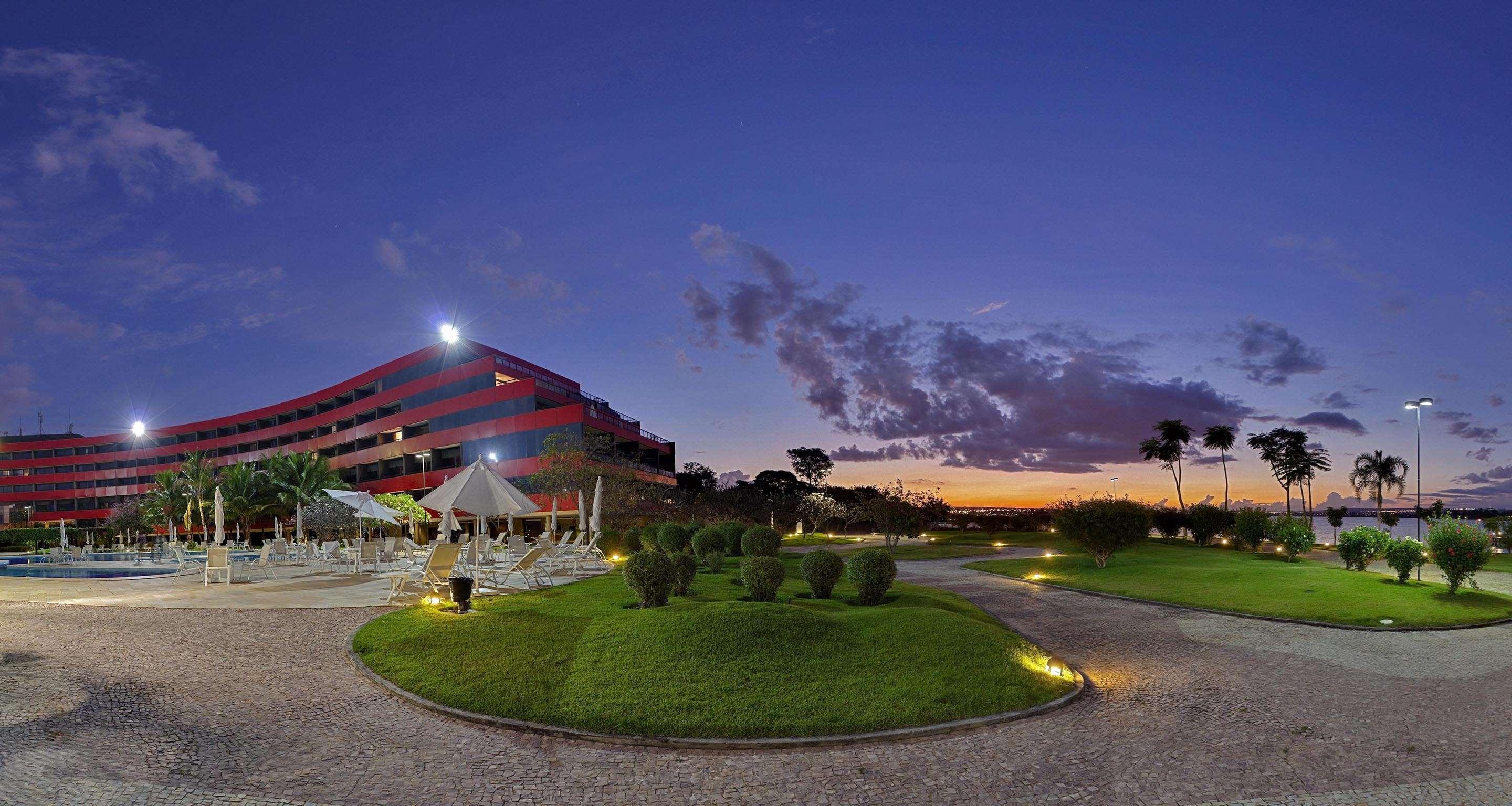 Royal Tulip Brasilia Alvorada Hotel Exterior foto The photo depicts a modern building, possibly a hotel or resort, with a striking red exterior. It is set in a landscaped area that features well-maintained greenery and ornamental plants. In the foreground, there are lounge areas with umbrellas, sugg