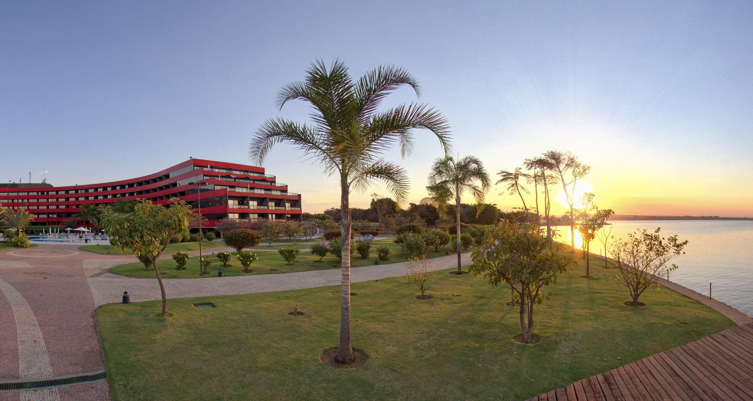 Royal Tulip Brasilia Alvorada Hotel Exterior foto The photo depicts a scenic view featuring a red multi-story building alongside a serene waterfront. In the foreground, there are several palm trees and lush green lawns, creating a tropical atmosphere. The sun is setting in the background, casting wa