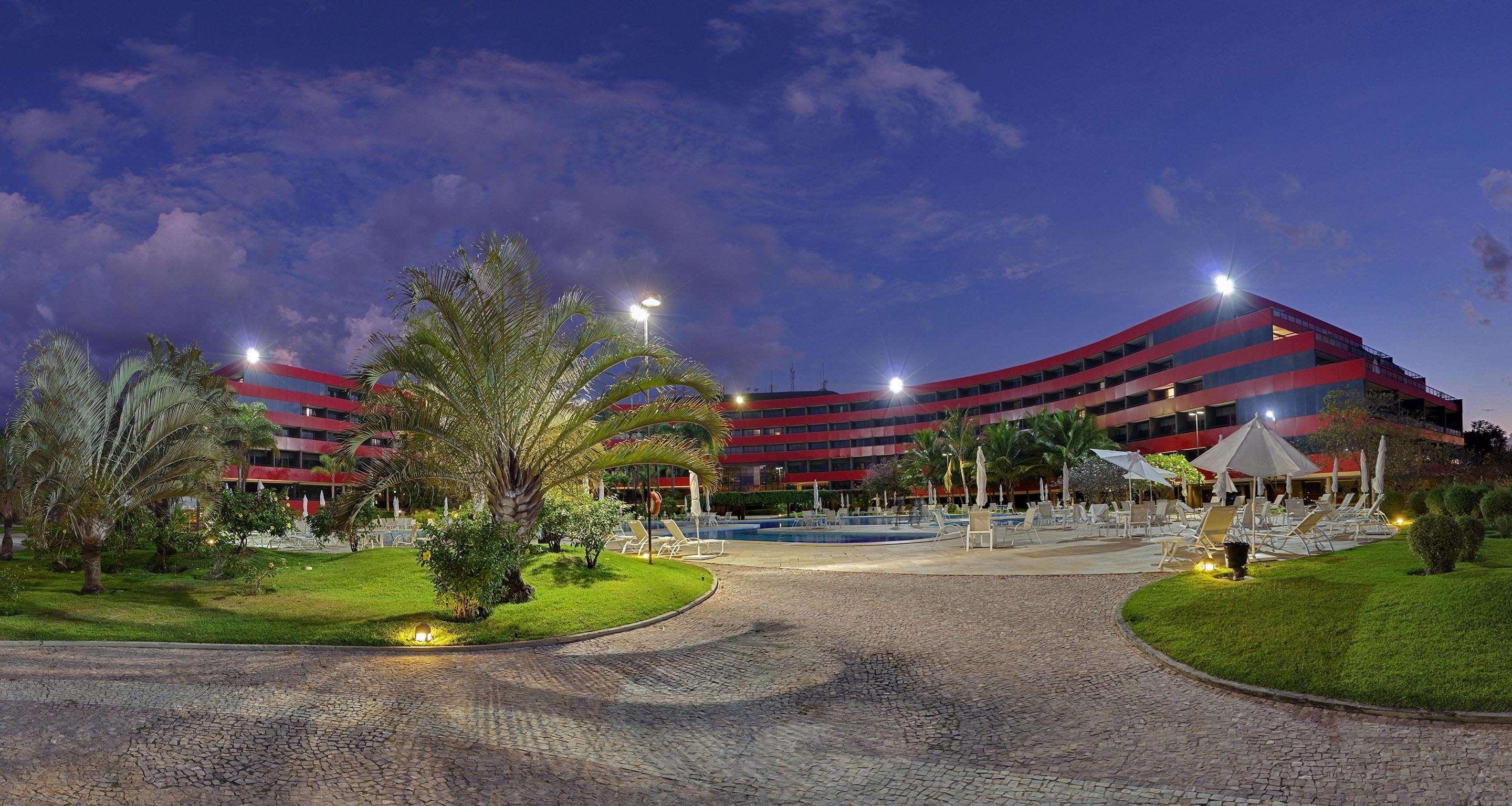 Royal Tulip Brasilia Alvorada Hotel Exterior foto The photo shows a panoramic view of a hotel or resort complex, characterized by its modern architectural design with smooth, curved lines and a vibrant red color. It’s set in a landscaped area featuring palm trees and green grass. The surroundings in