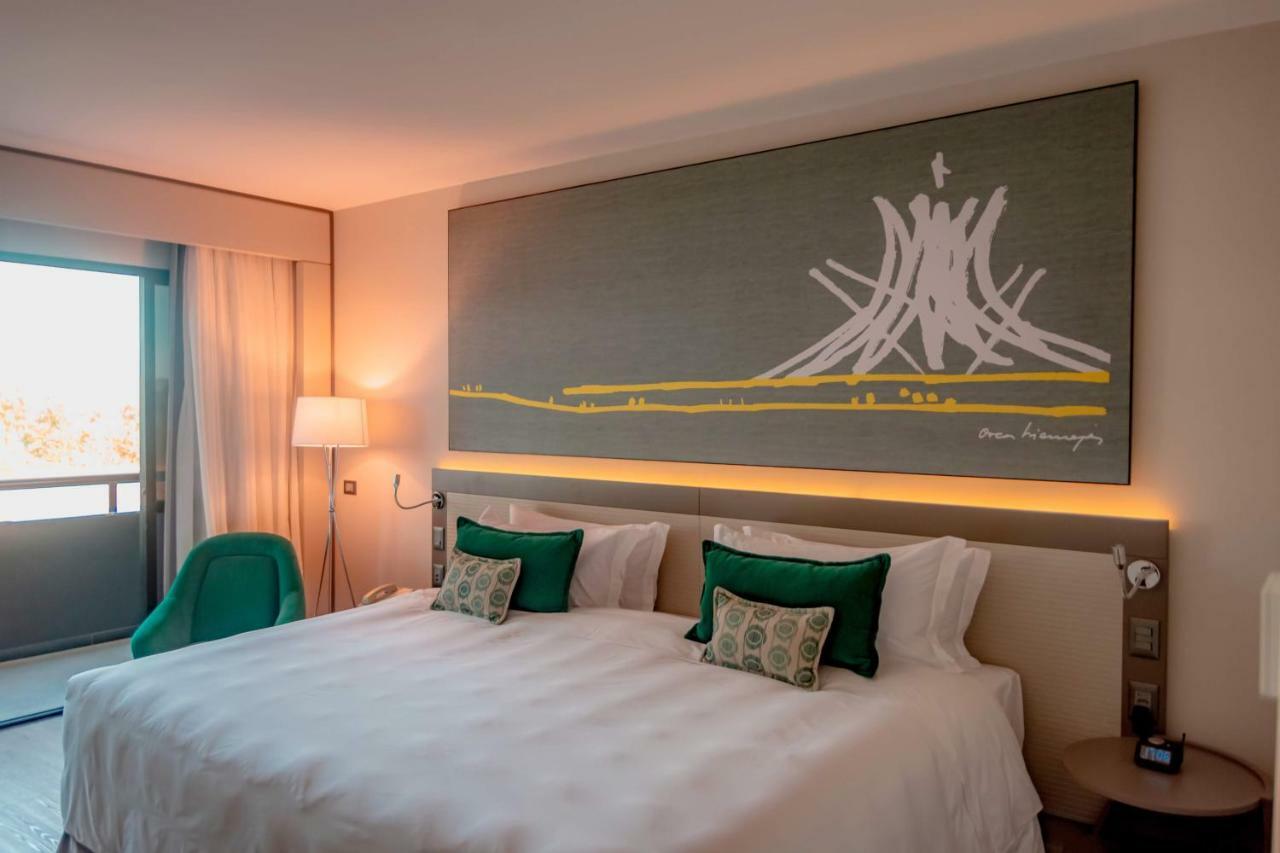 Royal Tulip Brasilia Alvorada Hotel Exterior foto The photo shows a modern hotel room featuring a large bed with white bedding and two green accent pillows. Above the bed, there is a large piece of artwork depicting a stylized mountain or volcanic shape with yellow accents. The room has a contempora